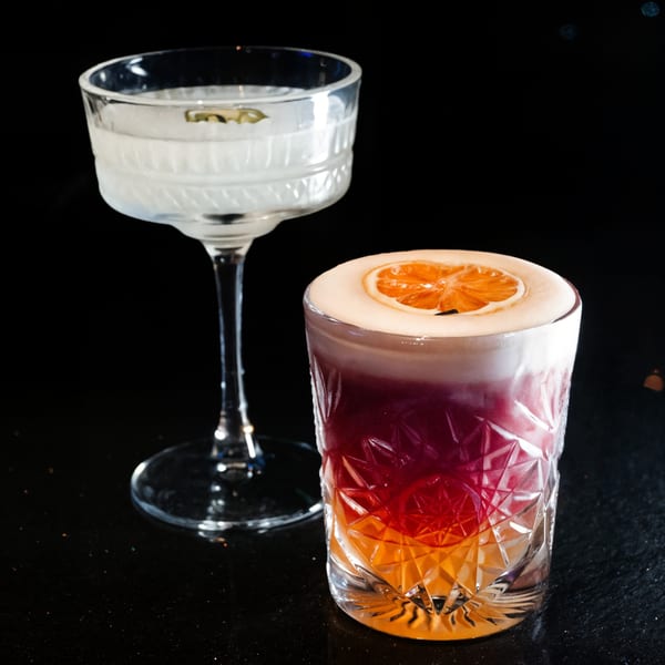 Two cocktails, one clear in a coupe glass and one layered red/orange with foam and orange slice.