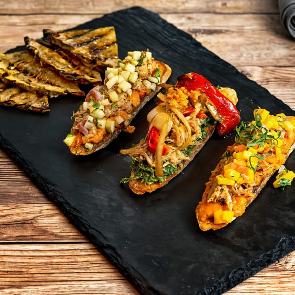 Three gourmet bruschettas with various toppings and grilled bread on a black slate board.